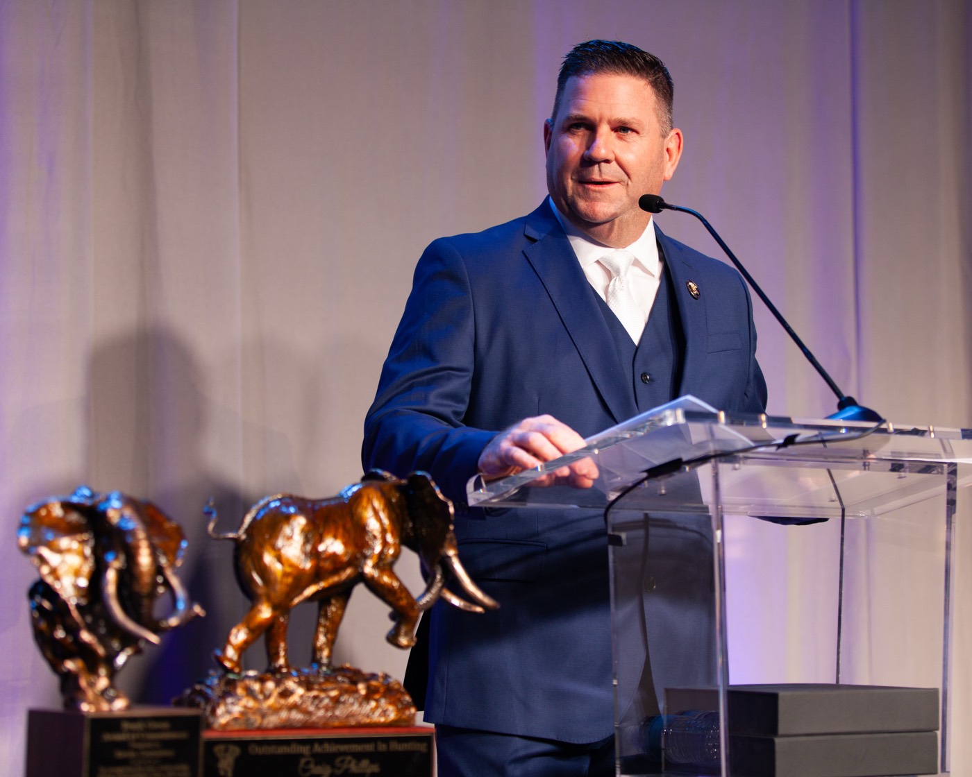 A speaker addressing a conservation banquet beside bronze wildlife trophies