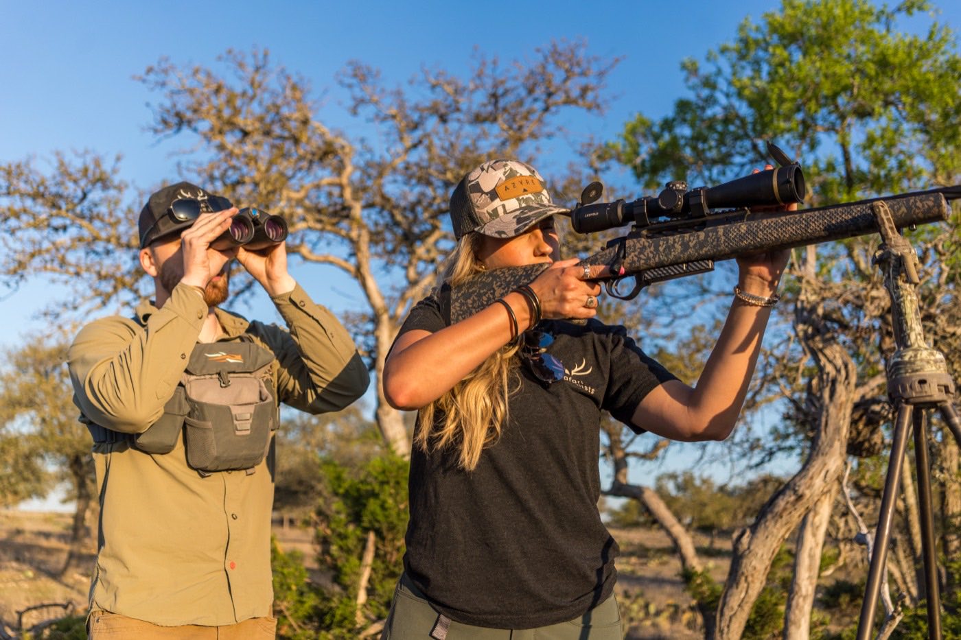 Two hunters glassing and taking aim from the edge of a treeline